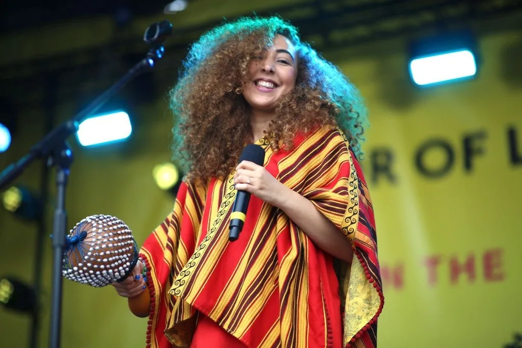 A singer smiling on an outdoor stage, holding a microphone and a frame drum
