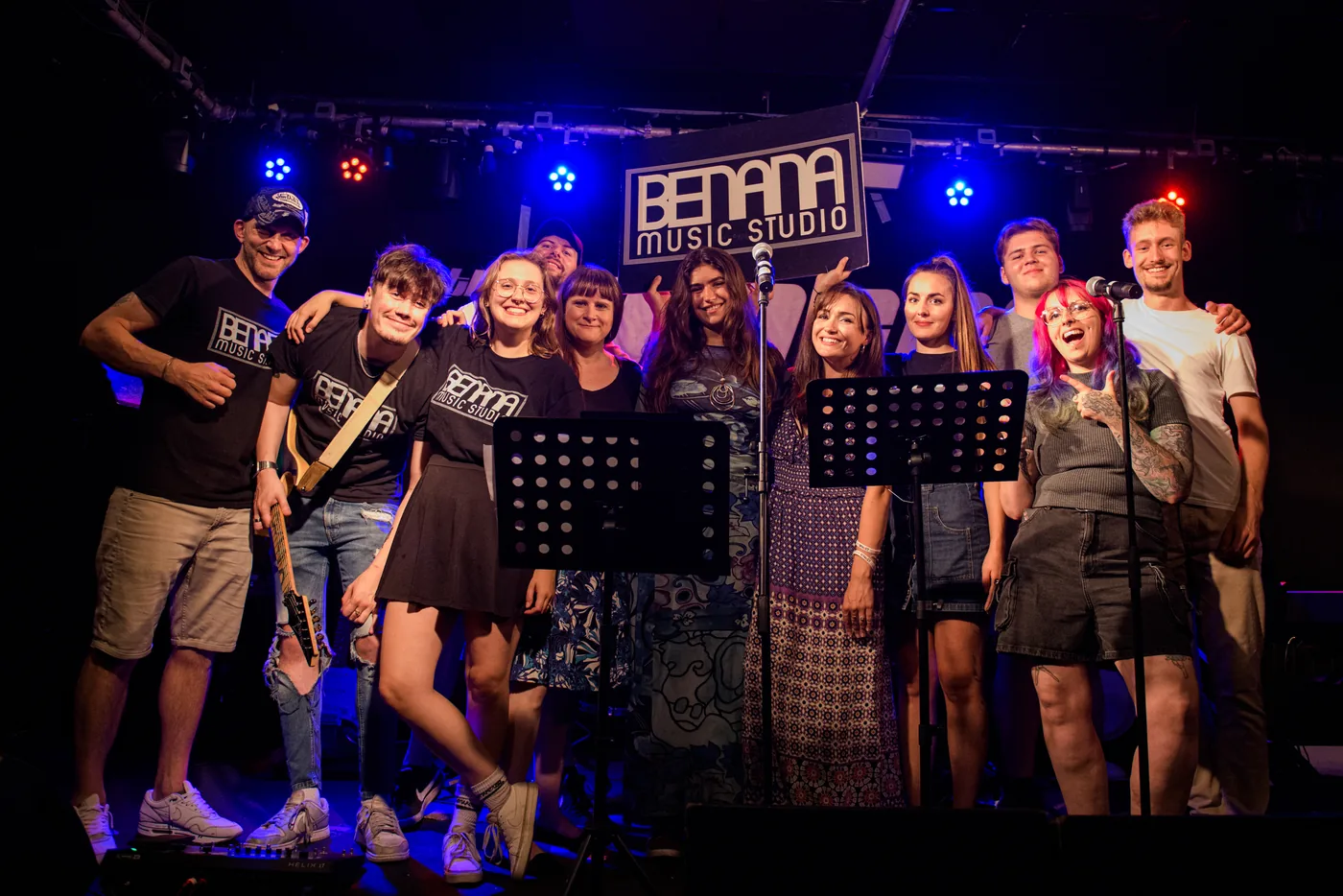 A large group of people of different ages standing together on a small stage under a Benana Music Studio banner, some holding instruments